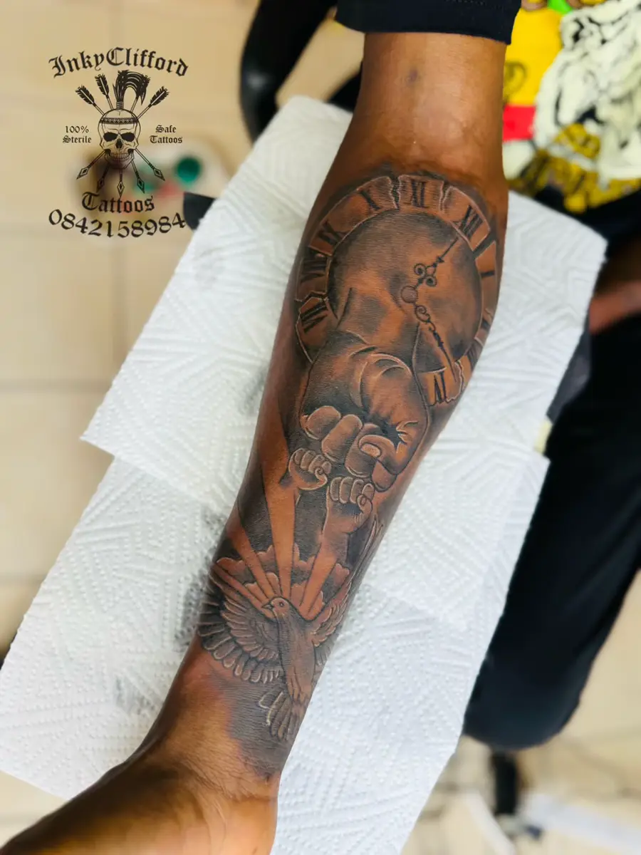 Forearm displaying a large black-and-gray tattoo sleeve with a clock, winged figure, and bird imagery