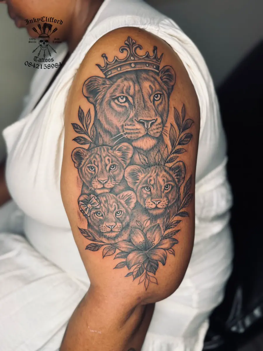 Upper arm tattoo featuring a crowned adult lion surrounded by three lion cubs, leaves, and flowers