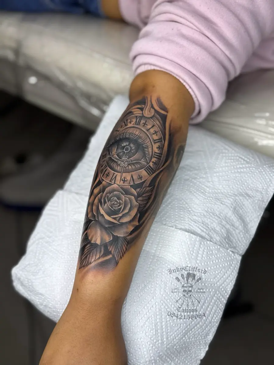 Forearm with a detailed black-and-gray tattoo featuring a compass-like clock design, rose, and leaves