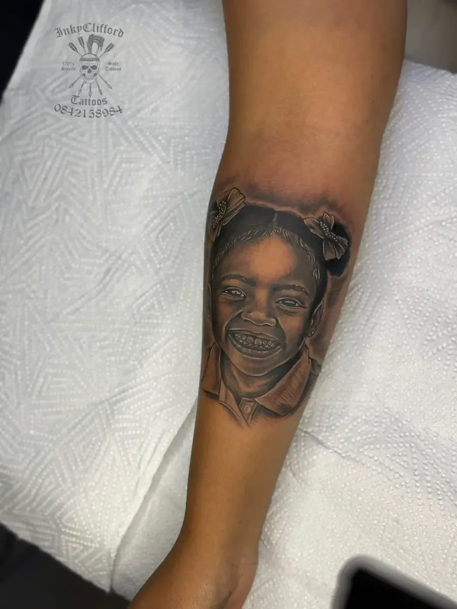 Detailed portrait tattoo of a smiling child with hair bows on a person's forearm over a white textured towel