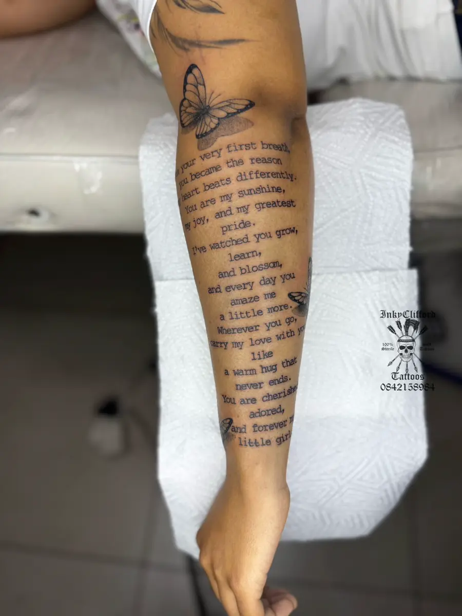 Tattooed forearm with a butterfly tattoo and stacked poem text resting on a white padded surface
