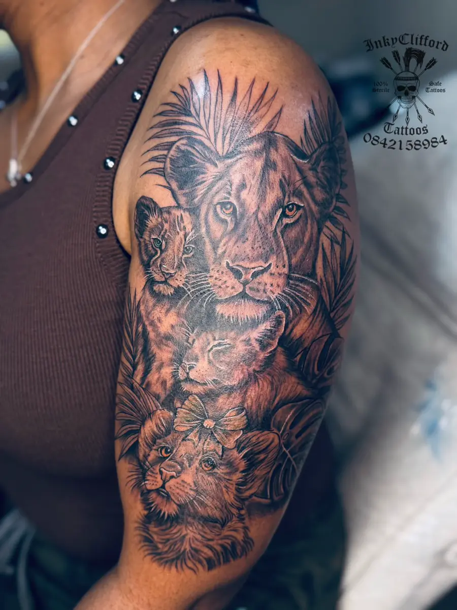 Upper arm featuring a detailed black-and-gray tattoo of a lion surrounded by several lion cubs and foliage